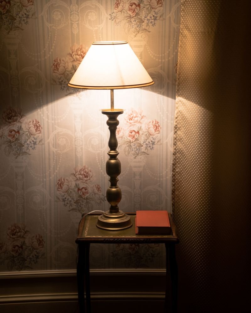 A turned brass table lamp with a pleated shade glowing against patterned wallpaper